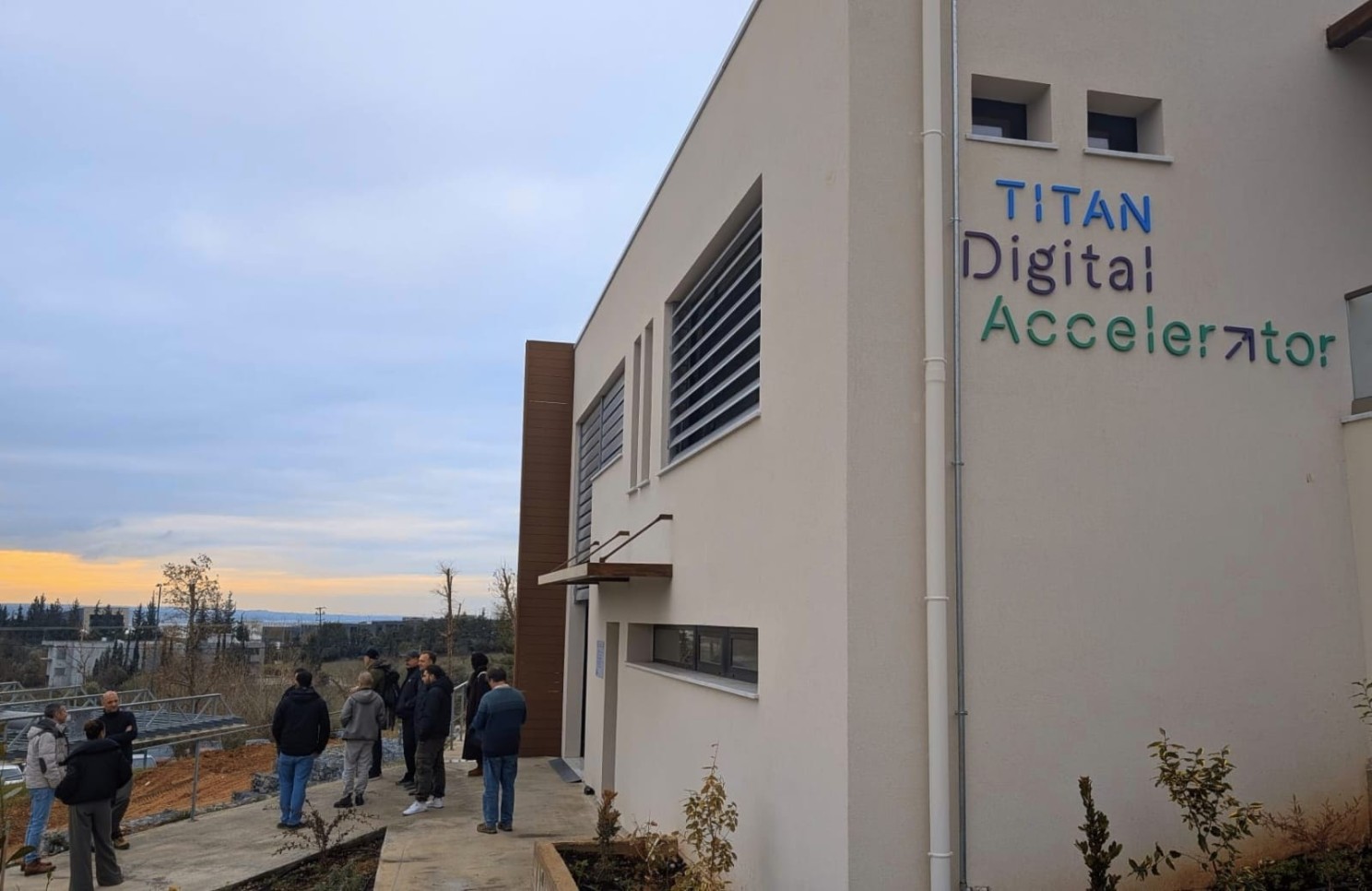 A group of people stands outside a modern building labelled "Titan Digital Accelerator" with a cloudy sky and distant trees in the background. A group of people stands outside a modern building labelled "Titan Digital Accelerator" with a cloudy sky and distant trees in the background.